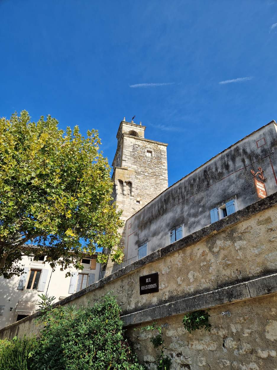 Le beffroi de Grignan, tour carrée en pierre au-dessus d'un mur, sous un ciel bleu